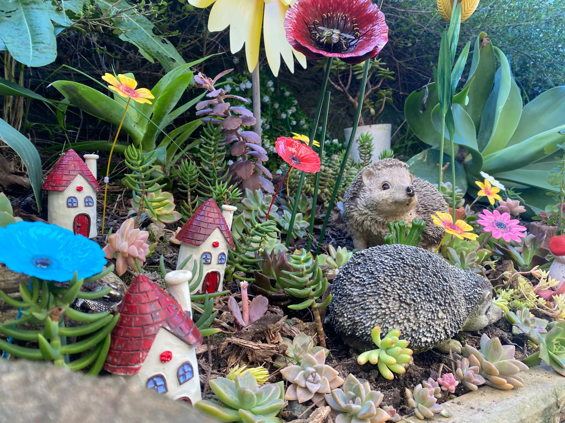 A fairy garden of tiny houses, succulents and ceramic flowers tucked into a garden bed