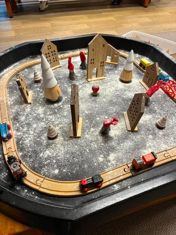 A wooden train track winter scene with timber houses, trees and peg dolls in a tuff tray