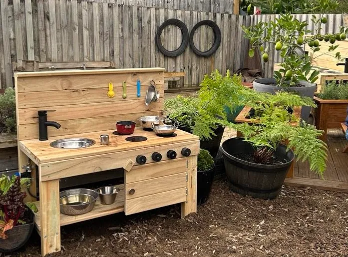 Outdoor mud kitchen with a sink, pots and pans surrounded by ferns and a lemon tree