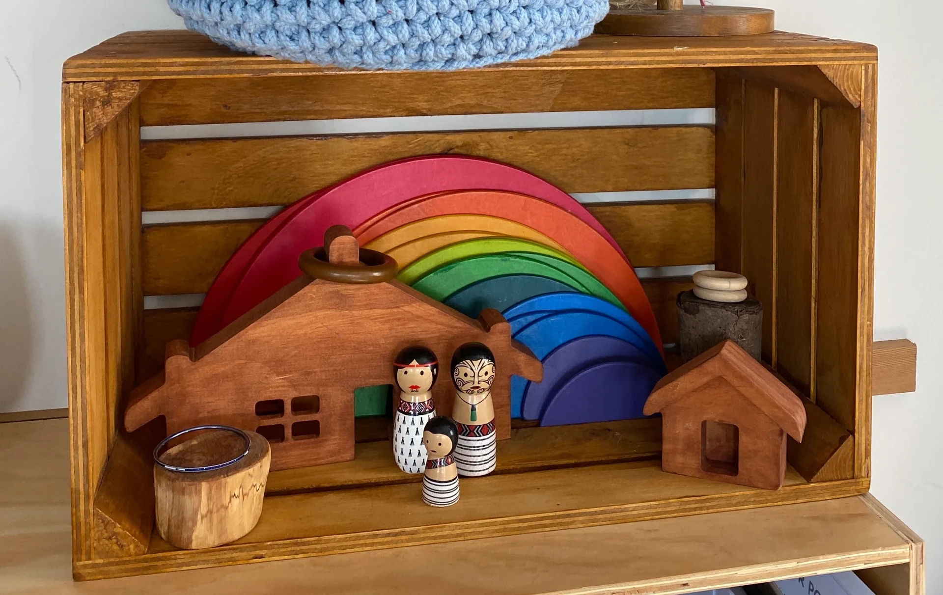 Wooden rainbow, house blocks and Māori peg dolls arranged in a timber crate shelf