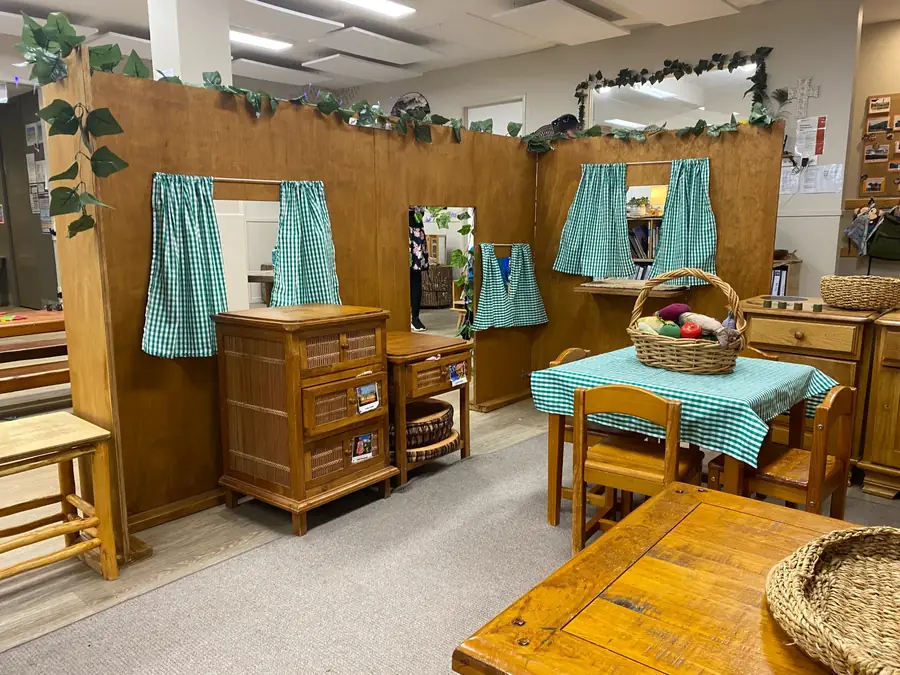 The Kākāpō Room home corner with a timber playhouse, gingham curtains and a set table